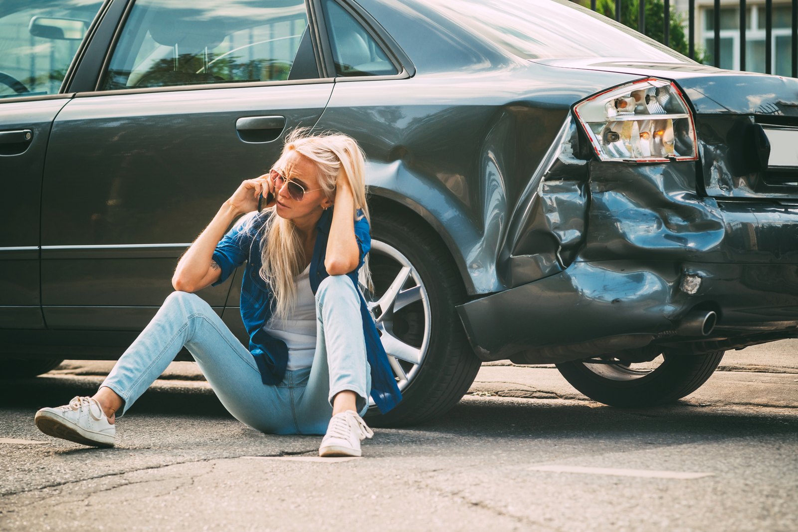 Woman sitting on the road beside a rear-ended car, on the phone after a crash in Texas