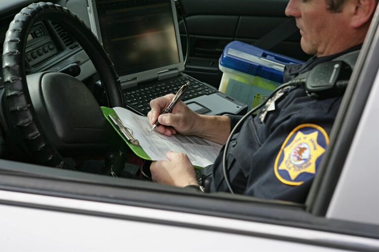 Police officer seated in a patrol car writing a crash report on a clipboard beside a mounted laptop