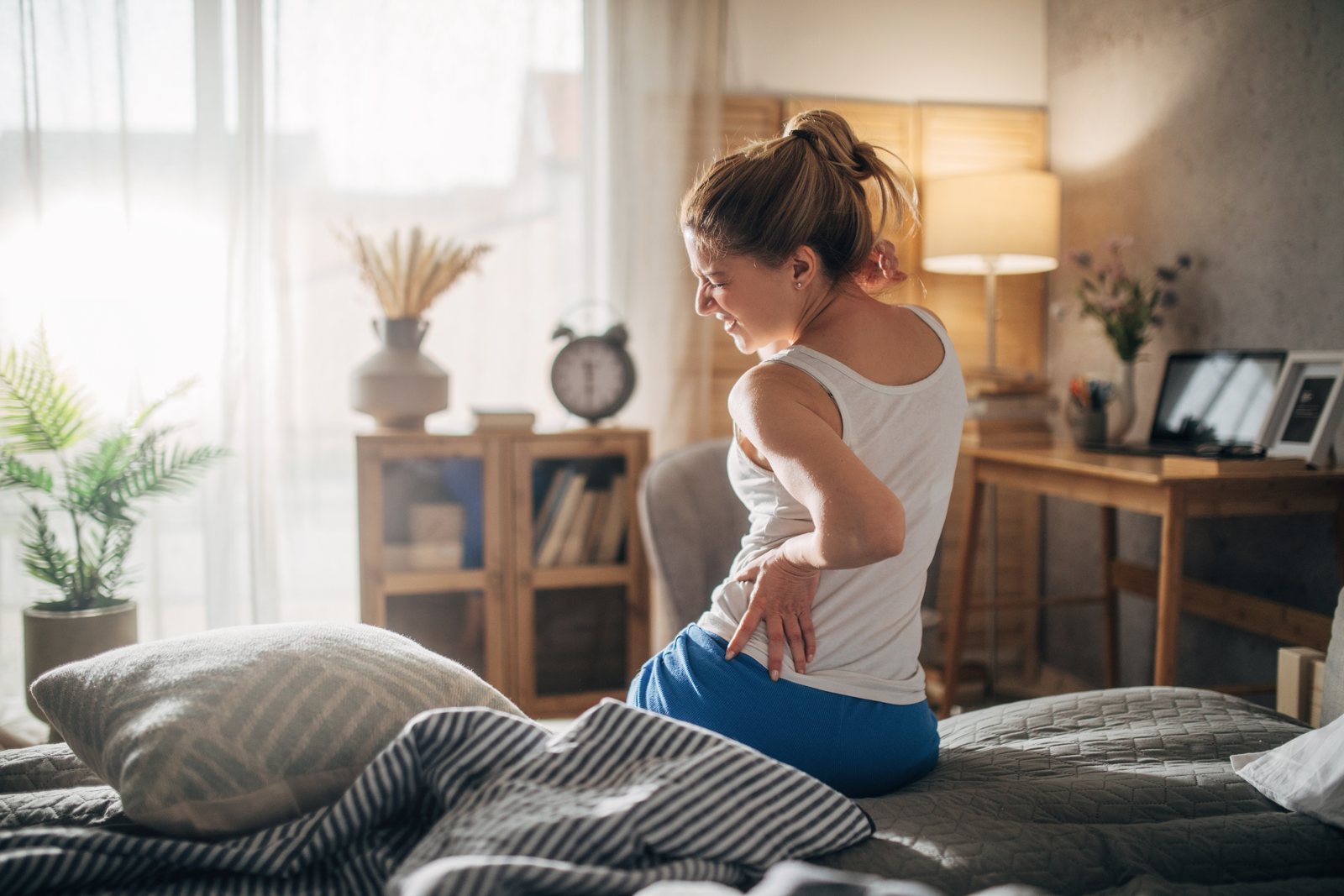 Woman sitting on the edge of her bed at home with a hand on her lower back, weeks after a car accident