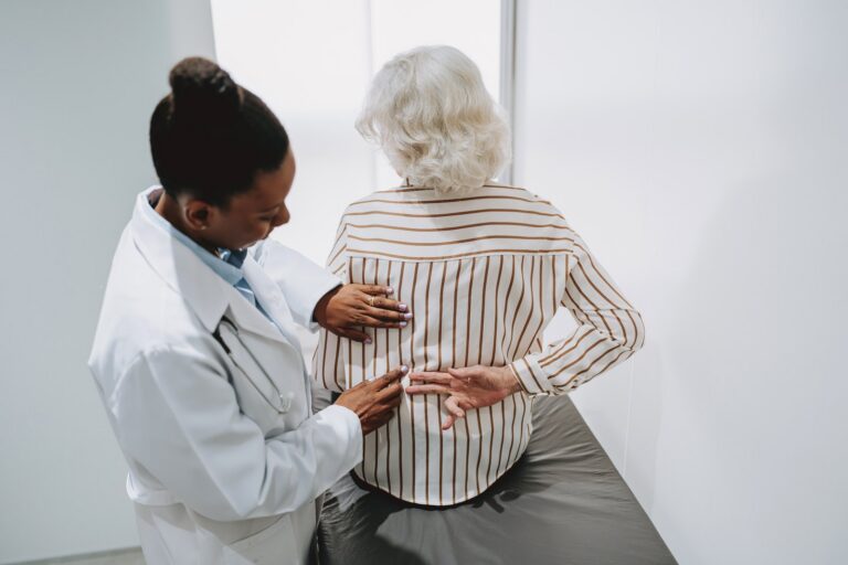 A clinician examining the lower back of an older patient seated on an exam table.