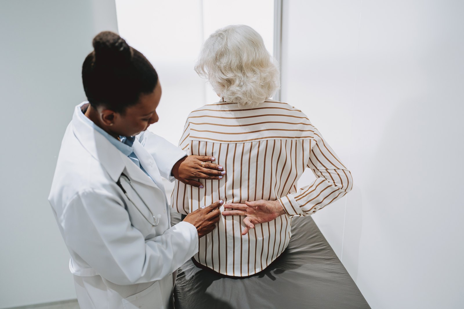 A clinician examining the lower back of an older patient seated on an exam table.
